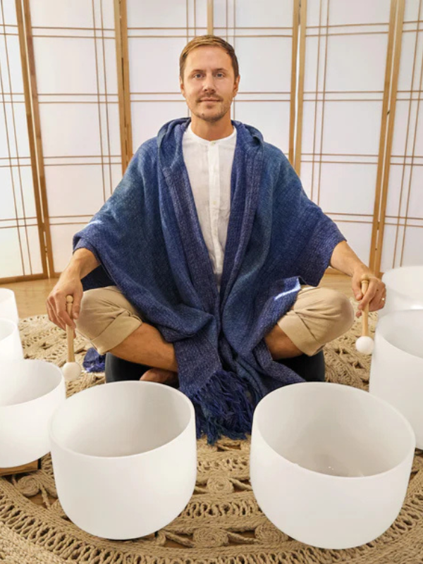 Man sitting in a meditative pose with crystal singing bowls around him, draped in a blue shawl.
